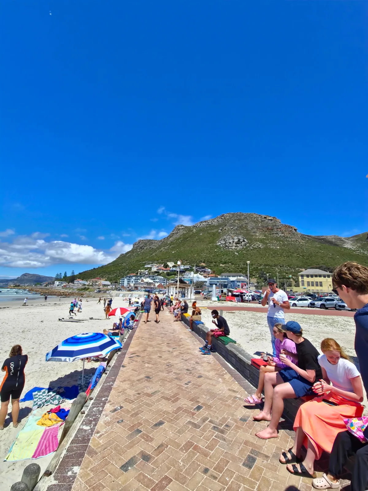 Muizenberg beachfront promenade with surfers, beachgoers and Muizenberg Peak rising above the False Bay coastline