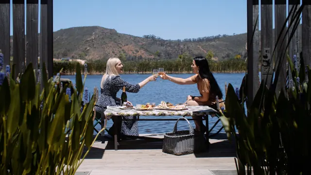 Two guests toasting at a picnic table on a deck above the water at Botlierskop