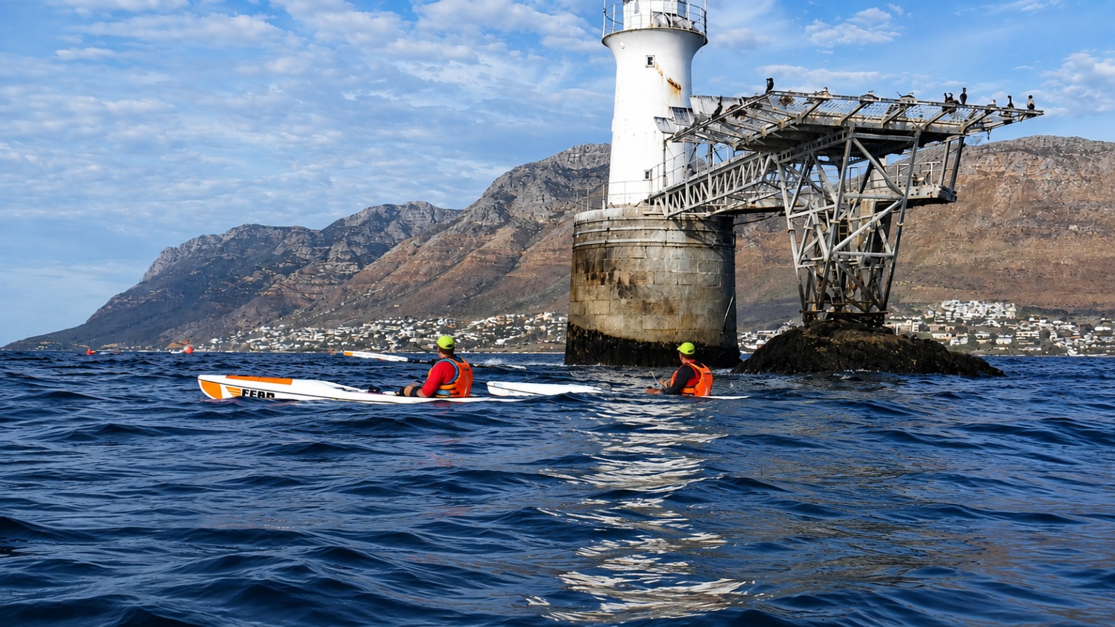 Surfski paddlers beside a False Bay lighthouse with mountains beyond